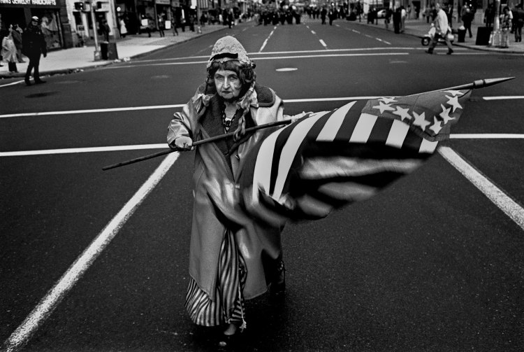 Flag Lady, NYC © Robert Goldstein. Black and white - elderly woman waving large stars and stripes flag in the street