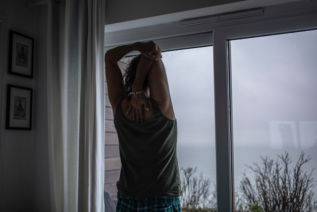 (self) Isolation - image © Jason Florio - a lone woman stretches in the early morning, looking out the window at the sea, Cornwall, UK
