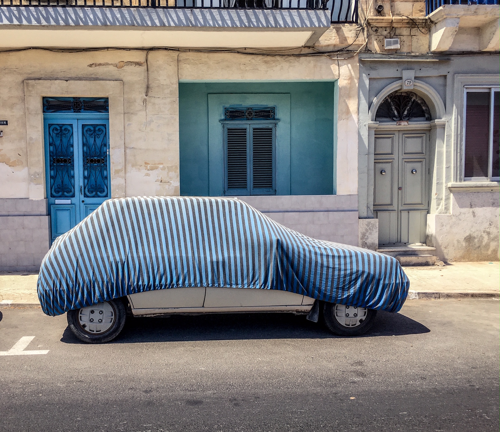 A car with blue striped cover, against blue house and doors, Malta. Image © Helen Jones-Florio