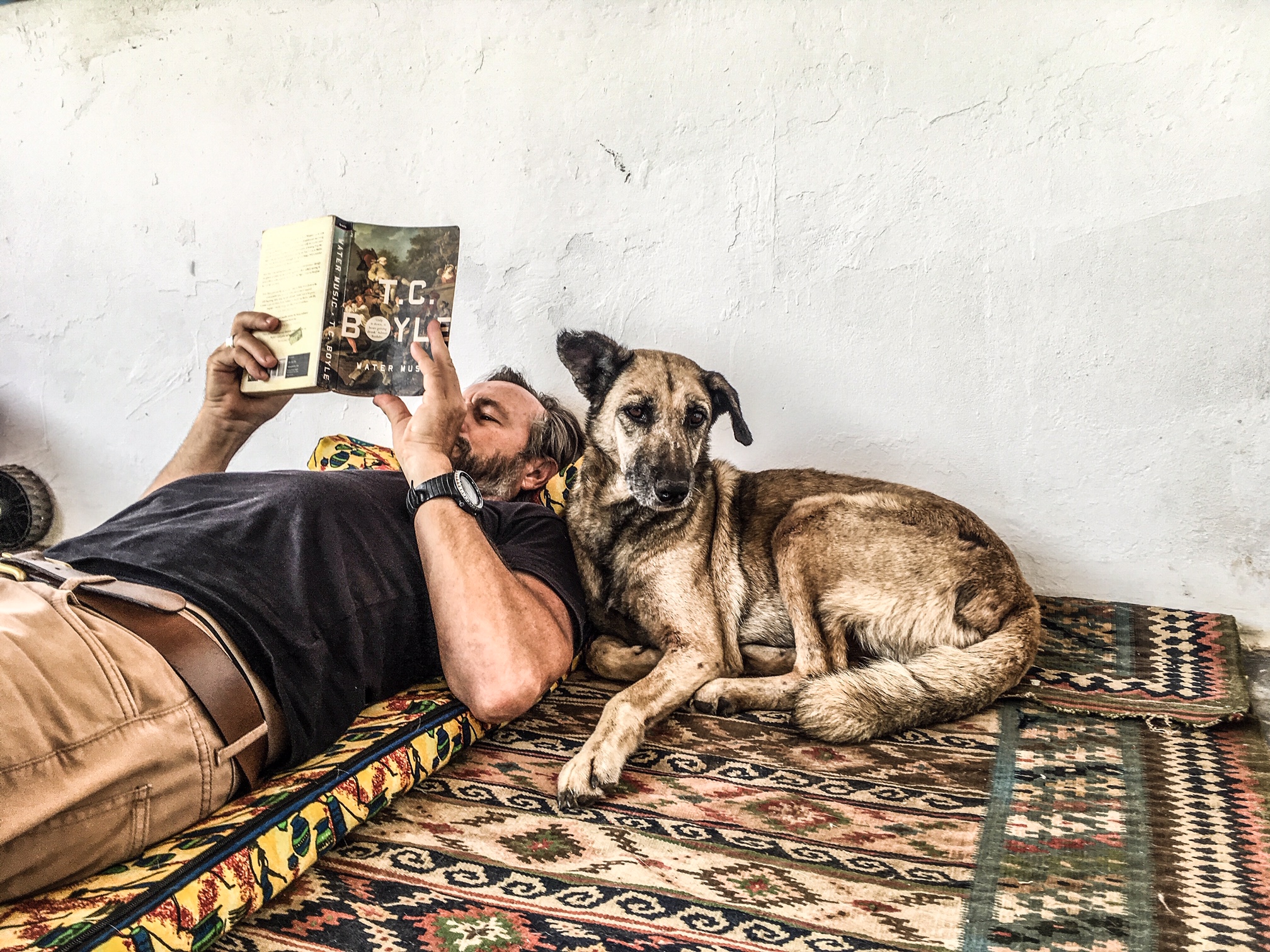Gambia dogs - a man reads a book, lying on a Persian carpet, with a dog lying next to him. Image © Helen Jones-Florio