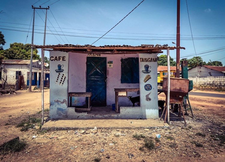 Gambia Doors: a local cafe storefront, with hand painted signs - with a goat hiding in the shade of the terrace - in a suburban setting. Image ©Helen Jones-Florio