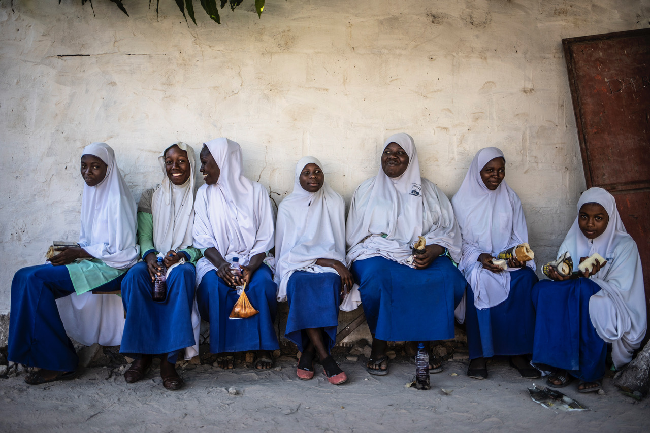 A group of Koranic school girls sitting together on a bench against a wall, smiling and laughing, The Gambia, West Africa. Image ©Helen Jones-Florio