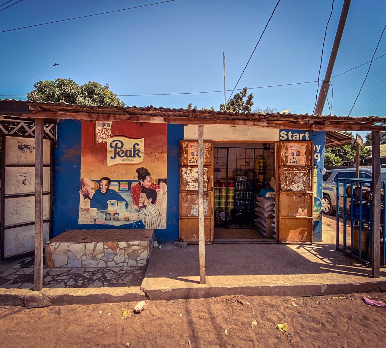 #GambiaDoors - a local shop, with a hand painted frontage, with a mural of a family sitting around the breakfast table, advertising 'Peak Milk', Bakau, The Gambia. Image ©Helen Jones-Florio
