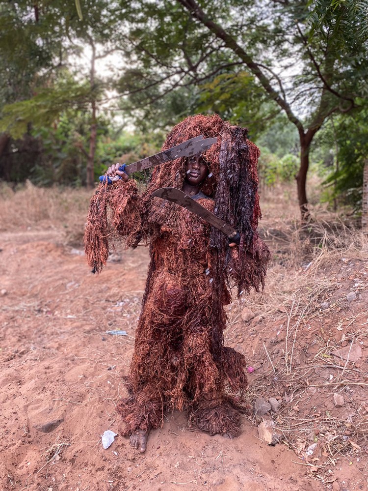 A young boy dressed up as a Kankurang - one of many traditional masquerades - holding two machete's standing amongst trees, The Gambia, West Africa. Image ©Jason Florio