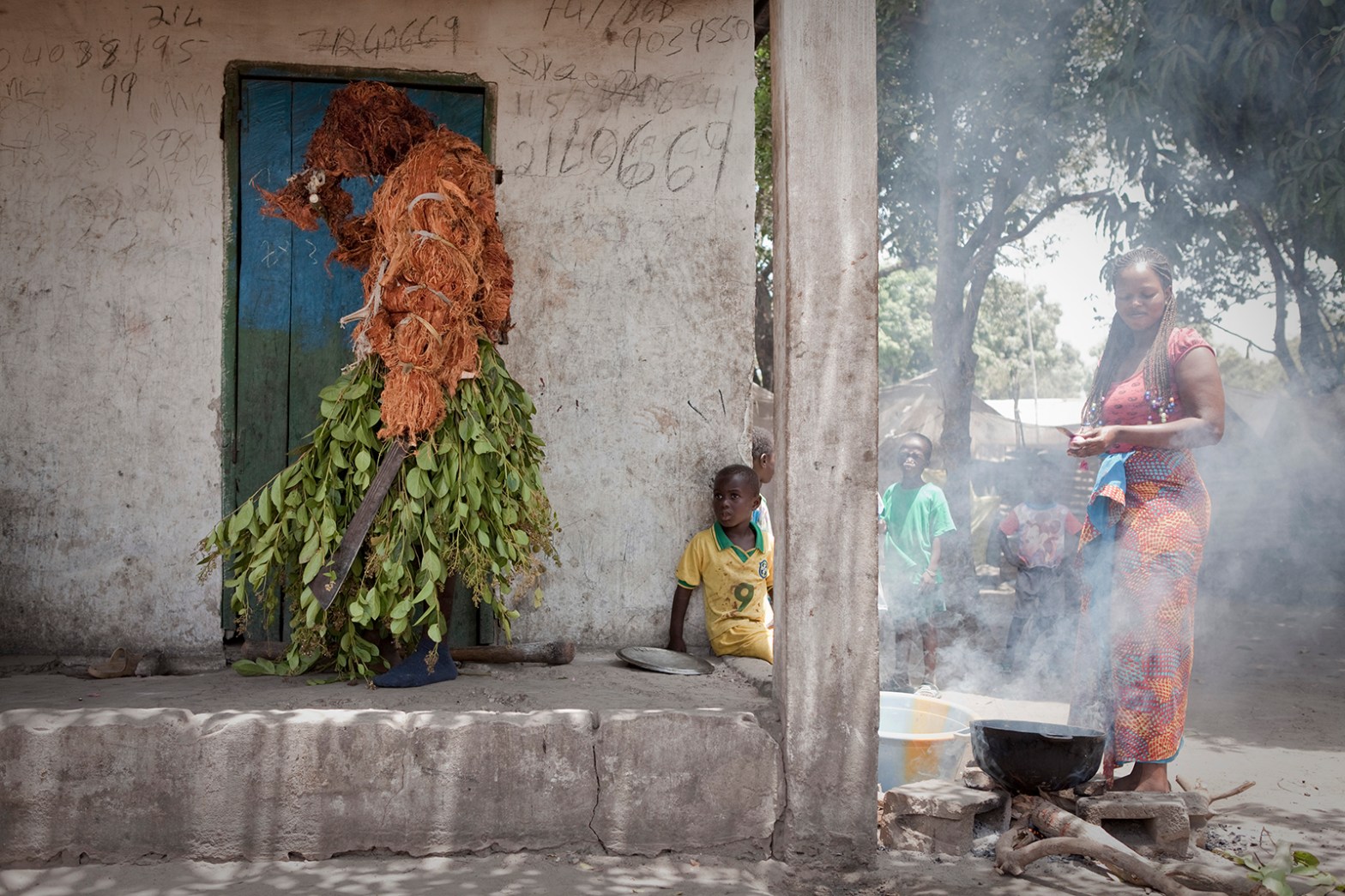 The Faara Kankurang, a tradition masquerade holding machetes, stands on the porch of a hut whilst a small child looks at him, and a woman cooks food over an open fire. The Gambia, West Africa. Image ©Jason Florio