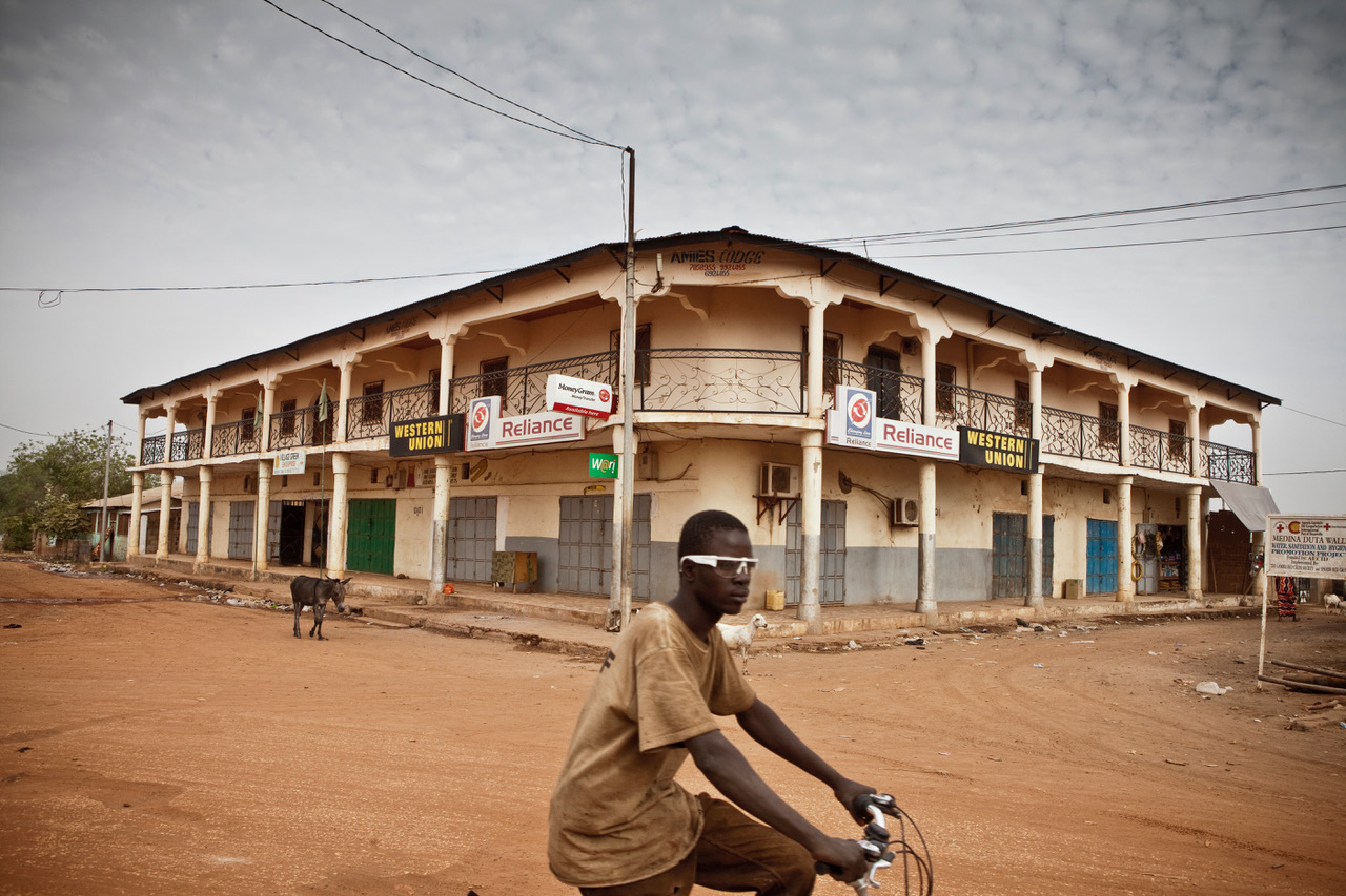 Photo fo the Day: A young boy wearing sunglasses, riding his bike in the town of Bansang, with a donkey in the background. Image ©Jason Florio